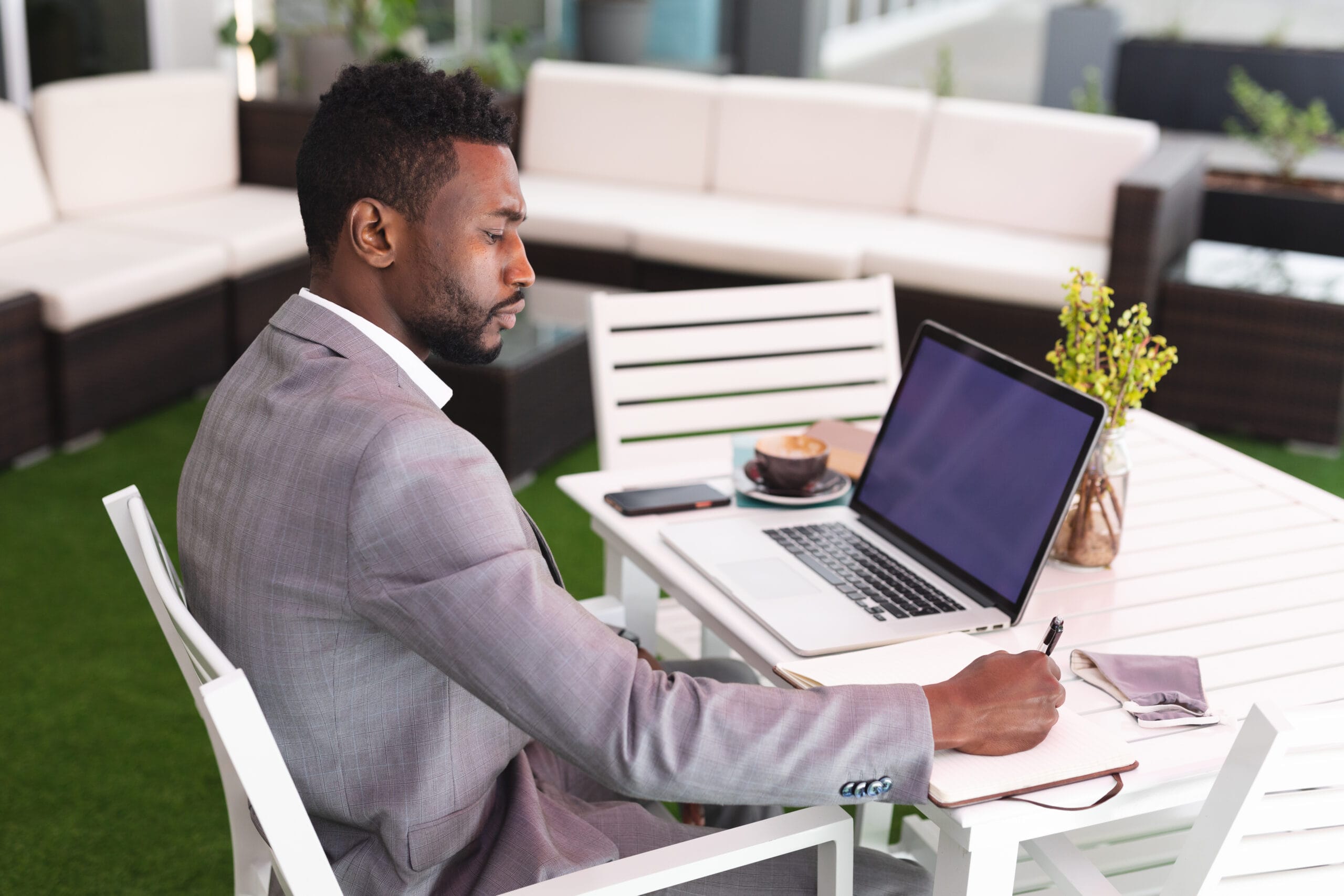 Man in a light suit working on a laptop and writing notes at an outdoor table, representing real estate business tasks.