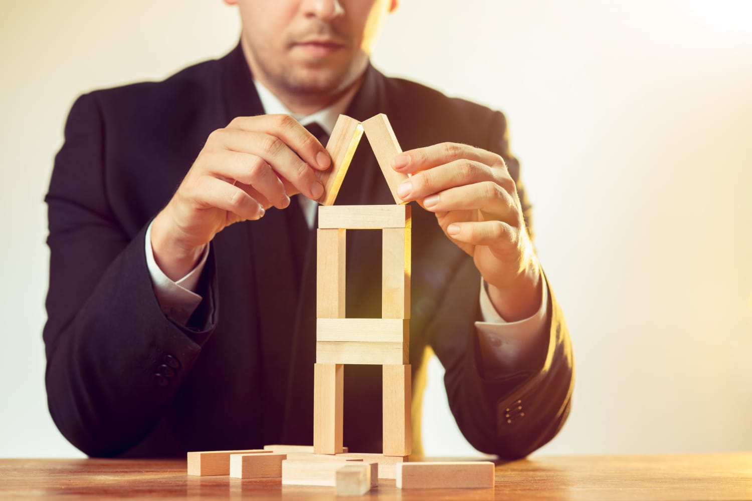 Man in a suit building a small wooden house model with blocks on a table