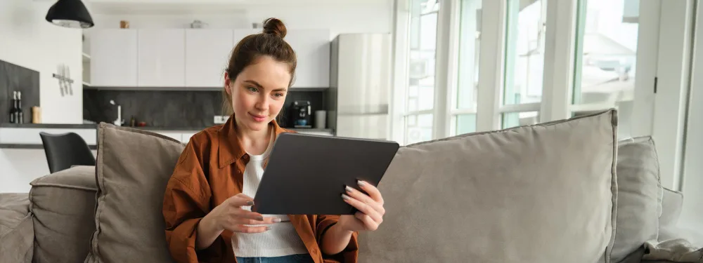 Young woman sitting on a couch, browsing real estate listings on a tablet.