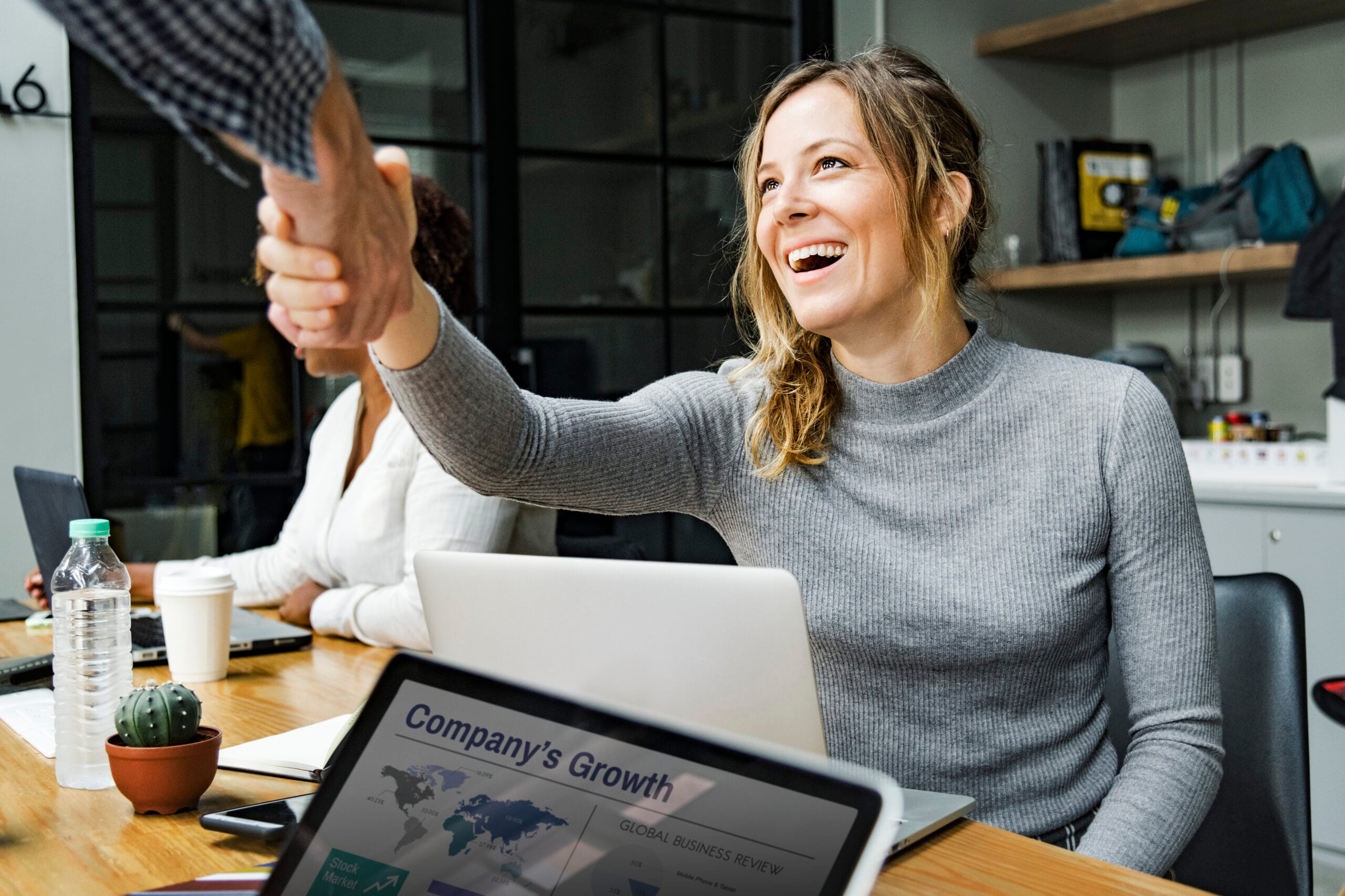 Happy woman celebrating success with handshake in modern office while laptop displays company growth analytics dashboard, representing successful SaaS affiliate partnership achievements